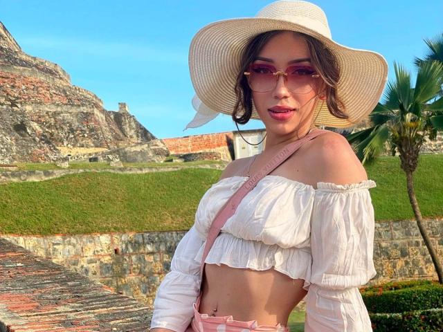 LindaHotx con gafas de sol rosas, una blusa blanca con un hombro descubierto, una correa rosa, un sombrero blanco, cabello castaño y una sonrisa encantadora, mostrando sus pechos y un piercing rosa en el pezón..