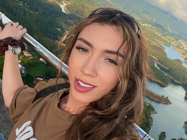 LindaHotx, com cabelo castanho ondulado, olhos verdes e lábios rosados, segurando uma barra de metal, sorrindo com a língua de fora, vestindo uma camisa marrom e uma faixa de cabelo no pulso..