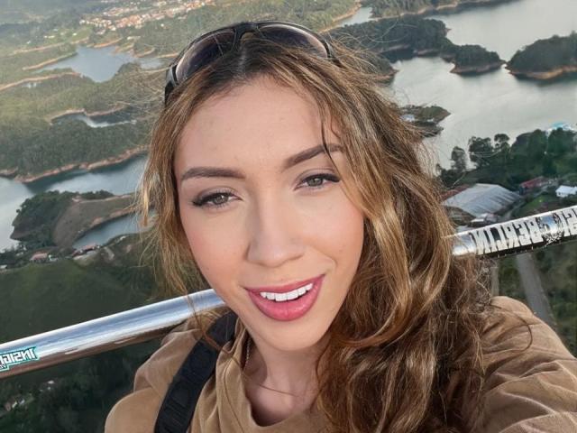 LindaHotx con cabello castaño ondulado, con gafas de sol en la cabeza, sonriendo con lápiz labial rosa, sosteniendo un tubo de metal plateado, vistiendo una camisa marrón, dientes blancos visibles, ojos verdes..