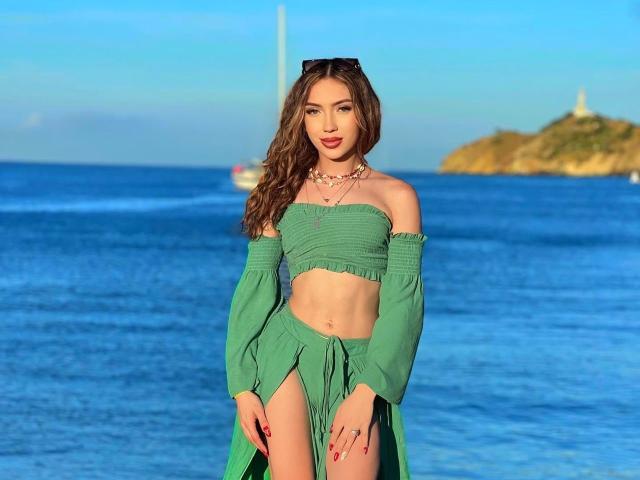 LindaHotx usando uma blusa cropped verde e uma saia combinando, mostrando seu abdômen tonificado e ombros descobertos, em uma praia com água azul e um barco ao fundo..