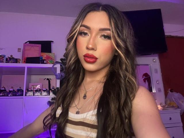 LindaHotx has wavy brown hair with blonde highlights, red lipstick, and wears a striped tank top, showing off her tits. She's looking at the camera with a sexy smile.
