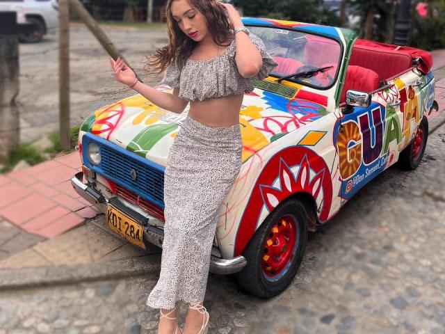 LindaHotx steht neben einem farbenfrohen, bemalten Auto, trägt ein graues, schulterfreies Crop-Top und einen passenden, hoch taillierten Rock, hält einen Holzstab und hat welliges, braunes Haar..