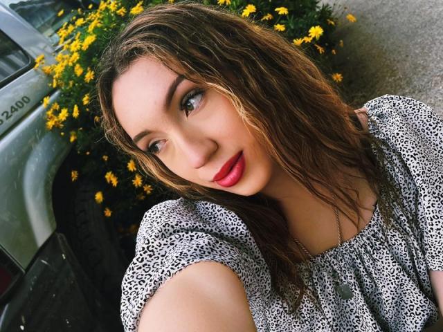 LindaHotx tem cabelos castanhos e molhados, olhos azuis, batom vermelho e está vestindo uma camisa com estampa de leopardo. Ela está encostada em um carro com flores amarelas. Seus seios são pequenos..