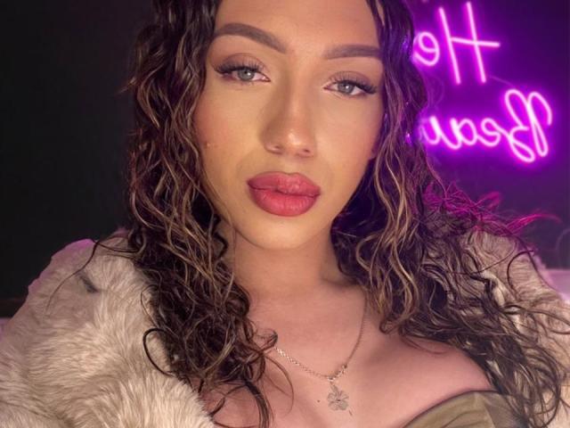 LindaHotx has wavy brown hair, full pink lips, green eyes, and wears a furry white jacket. Her tits are partially visible, and she's wearing a silver necklace.