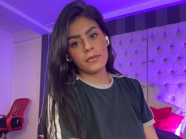 CamilePretty, with long black hair, wearing a green and white Adidas shirt, has a cute face and a small nose, looking at the camera with a subtle smile.