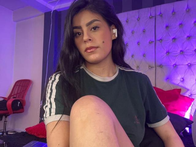 CamilePretty, a young woman with long black hair and medium brown skin, sits with a serious expression. She wears a green and white Adidas sports shirt, with a small logo on the chest. Her right knee is up, partially covering her black shorts. She has a small beauty mark under her right eye. The background includes a tufted headboard with purple lighting, a red chair, and a bed with red pillows. She also wears white earbuds. The image has a casual, slightly messy bedroom setting.