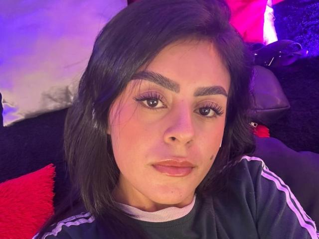 CamilePretty has dark brown hair, thick eyebrows, long eyelashes, and is wearing a green and white Adidas shirt. Her pink lips are slightly parted.