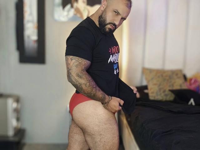 BardoCkroy69, bearded, bald, tattooed, wearing black shirt, red boyshorts, pulling shirt down, showing hairy butt, standing by bed, looking back, bedroom, serious expression.