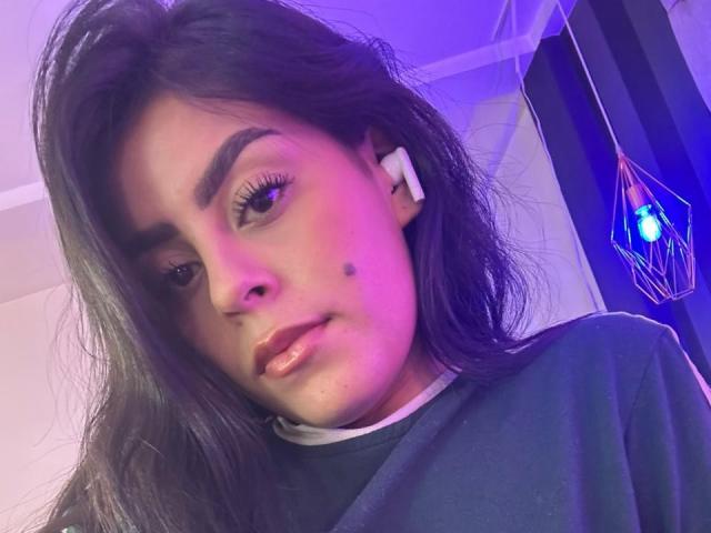 CamilePretty has long dark hair, a small mole on her right cheek, and is wearing white earbuds with a purple-blue light in the background. She's wearing a black shirt.