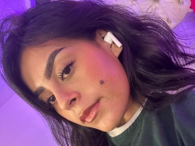 Close-up of CamilePretty with medium brown skin and long black hair. She has thick, well-groomed eyebrows and is wearing white earbuds in her right ear. A small black mole is visible on her right cheek. Her lips are slightly parted with a natural pinkish hue. She wears a dark green shirt with a white collar. The lighting has a purple tint, highlighting her cheek and forehead. Her expression is neutral with a slight smile. The texture of her hair is slightly wavy and loose.