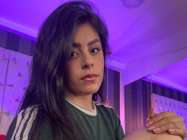 CamilePretty with long black hair, brown eyes, and full lips, wearing a green sports shirt with white stripes, holding her knee, under purple lighting.
