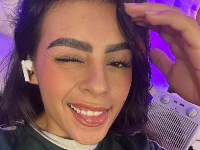 Close-up of CamilePretty with medium brown skin, dark eyebrows, and long black hair. She's winking, sticking out her tongue, and smiling with glossy pink lips. Her right hand is touching her hair. She's wearing white earphones and a green and white shirt. The background has purple lighting. Her eyelashes are long and she has a slight blush on her cheeks. The image has a casual, playful vibe.