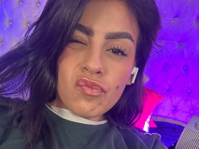 CamilePretty with shiny pink lips, winking, black hair, white earbuds, and a small beauty mark on her right cheek. She's making a pouty face, looking sexy.