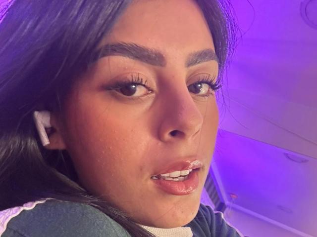 Close-up photograph of CamilePretty. She has long dark hair, thick eyebrows, and full lips with glossy pink lipstick. Her brown eyes are slightly open, and she's wearing white earbuds. Her skin is smooth with visible small pores. She's looking to the right, and the background is purple with a white ceiling. Her expression is neutral. The lighting is soft, highlighting her facial features.