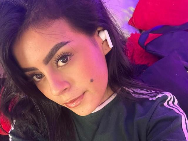 CamilePretty has medium brown skin, dark brown eyes, and black hair with a small mole on her right cheek. She's wearing white earbuds, a black Adidas shirt, and smiling.