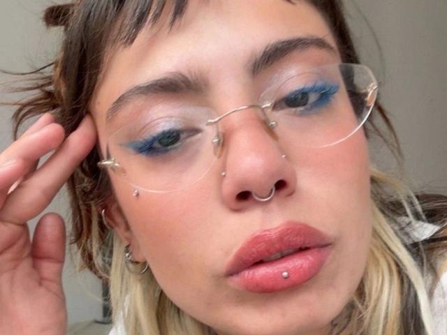 LunaRoses-hot has blue eyeshadow, clear glasses, nose piercings, lip piercing, and a nose ring. Her blonde-brown hair is messy, and she's touching her head with a hand.