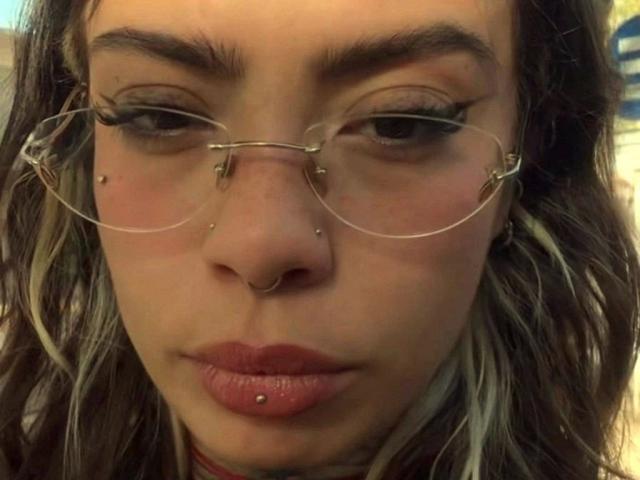 LunaRoses-hot has a nose ring, lip piercing, and clear glasses with metal clips. She's got a small beauty mark above her lip and pinkish-red lips.