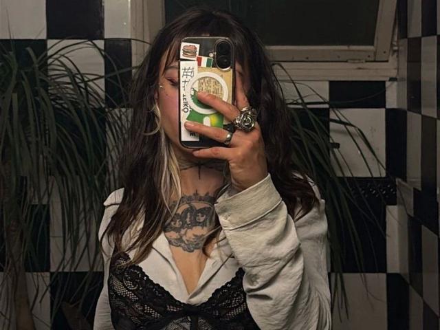 LunaRoses-hot with long dark hair, white-blonde highlights, lace top, white shirt, black tattoos, holding phone covering face, wearing rings, green phone case, black-white tiled wall background.