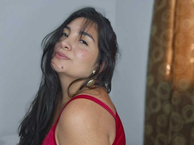CherrySweetX has dark, wavy hair, light skin, small moles on her cheek, and is wearing a red lace top. She's smiling slightly with a playful, cocky expression.