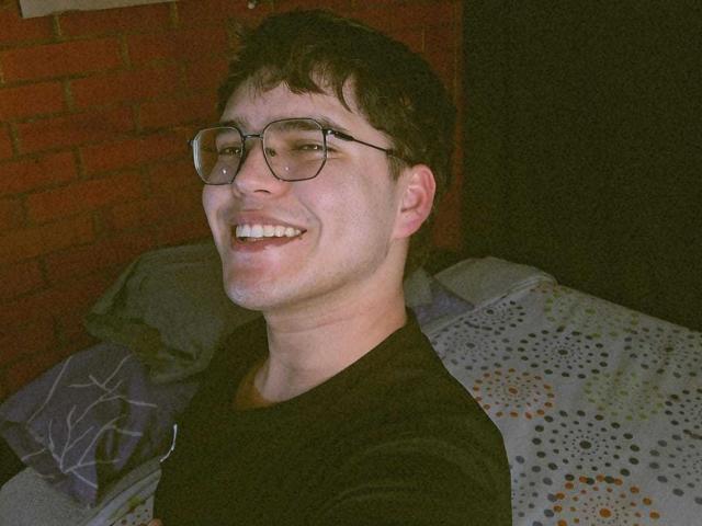 EddTerence is a nerdy dude with glasses, short brown hair, and a black shirt, smiling at the camera with a visible chin and nose. He's on a bed.