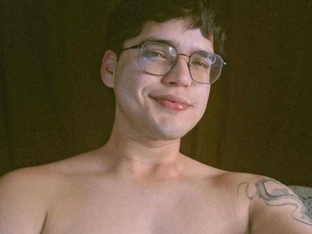 Shirtless EddTerence with glasses, short dark hair, and a tattoo on his right shoulder, smiling slightly. His nose is pierced. He's looking at the camera.