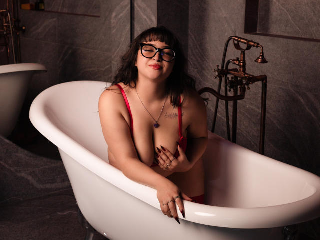 CherryMeloon with dark hair, glasses, and red lingerie in a white bathtub, covering her tits with one hand, smiling, tattoos on chest, brass faucet, dark marble walls.