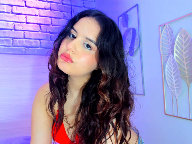 KylieLumi with wavy black hair, red lipstick, and a red tank top, looking slightly to the side with a subtle pout, in a blue-lit room.