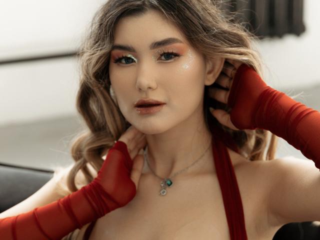 AnneTLuis has wavy blonde hair, red eyeliner, and red sheer finger gloves. She's wearing a red halter top, exposing her tits, with a teal necklace. She looks seductive.