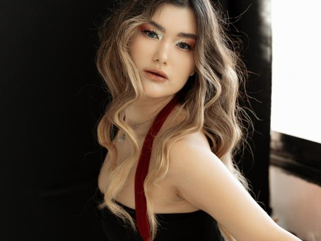 AnneTLuis with wavy blonde hair, red eyeliner, and subtle makeup, wearing black straps, has fair skin, and a necklace, looking at the camera with a slight pout.