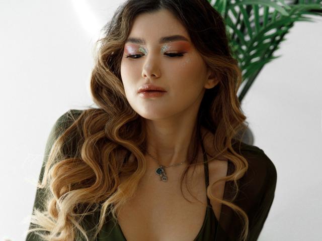 AnneTLuis has wavy, blonde-to-brown hair, glowing orange eyeshadow, and a green dress with a low neckline. She wears a silver necklace with a blue pendant.