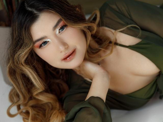 AnneTLuis has long, wavy brown hair with blonde highlights. She's wearing a sheer, green dress with a deep neckline, revealing part of her cleavage. Her eyes are blue with shimmering silver glitter, and she has full, pinkish lips. She's looking directly at the camera with a slight smile, her right hand touching her chin. The image is a close-up with soft lighting, highlighting her fair skin and delicate features. Her expression is relaxed and inviting.