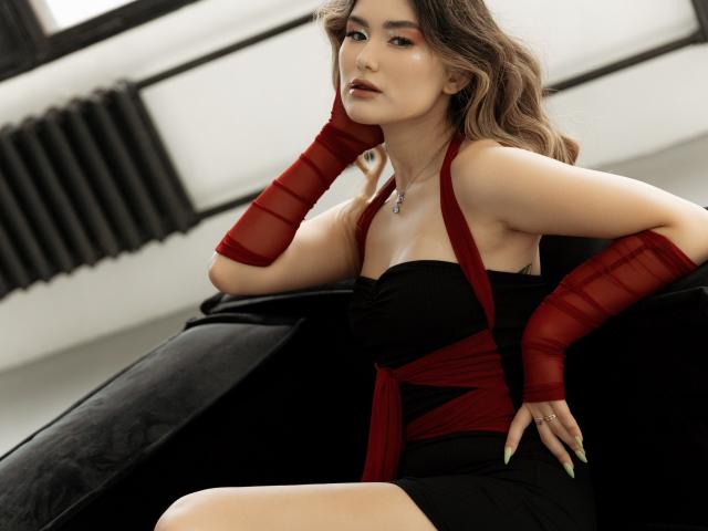 AnneTLuis with red mesh arm sleeves, black strapless dress, sitting on black couch, hand on cheek, looking at camera, pale skin, brown wavy hair, green nails, ring.