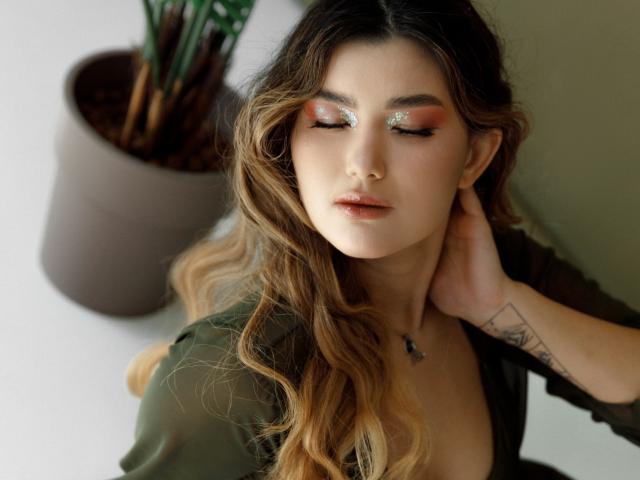 AnneTLuis with long wavy blonde hair, green eyes, and shimmering eyeshadow, wearing a green top, hand touching her neck, and a tattoo on her wrist, looking relaxed.