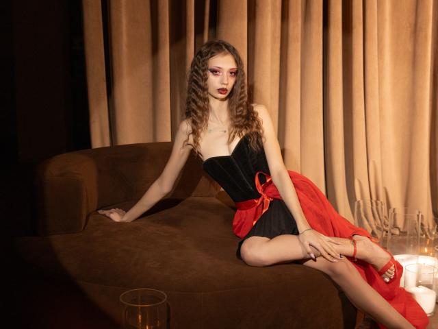 EmilyGraydis with curly brown hair, wearing a black strapless top and red skirt, sitting on a brown couch, legs crossed, red heels, dark red lipstick, looking to the side.