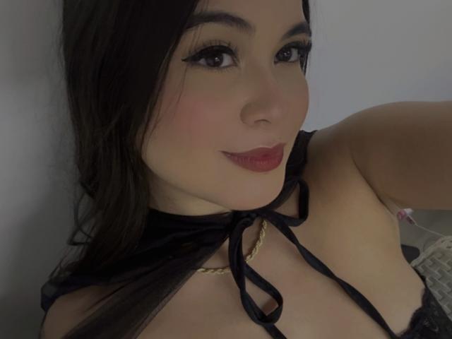 CamilaSofiia with long dark hair, wearing black lace top, revealing her boobs, has red lipstick, black eyeliner, and a gold necklace, smiling slightly.