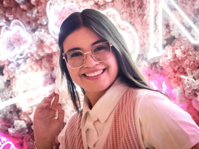 CamilaSofiia with long dark hair, glasses, smiling, wearing a pink houndstooth vest over a white shirt, holding her hair, pink floral background, cute, attractive.