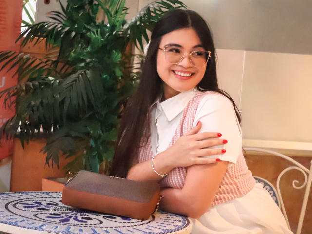 CamilaSofiia with long black hair, glasses, smiling, wearing a pink and white checkered dress, white shirt, red nails, brown purse on a blue-patterned table, arms crossed.