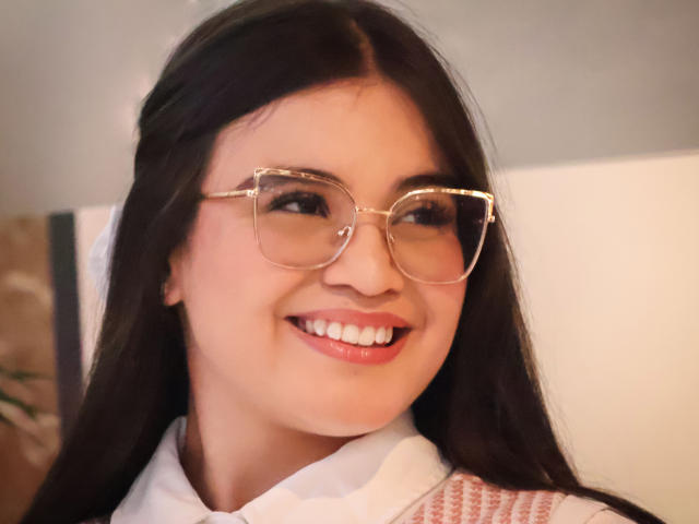 CamilaSofiia with long black hair, wearing gold-rimmed glasses, smiling with white teeth, and pink lipstick, looking cute with her ass-kissing smile. She's wearing a white shirt.