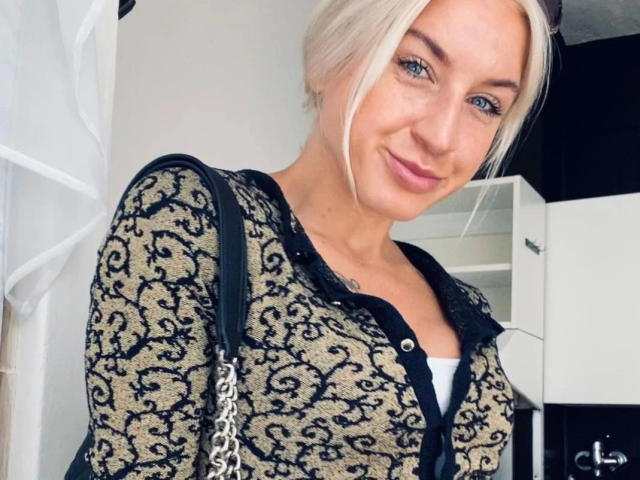 KayleenB-hot has blonde hair in a messy updo, blue eyes, and light skin. She wears a black and gold patterned cardigan over a white shirt. A black shoulder bag with a chain strap is on her right shoulder. She smiles softly. The image is a close-up, focusing on her upper body and face. The background is blurry. The composition centers on her face and upper torso.