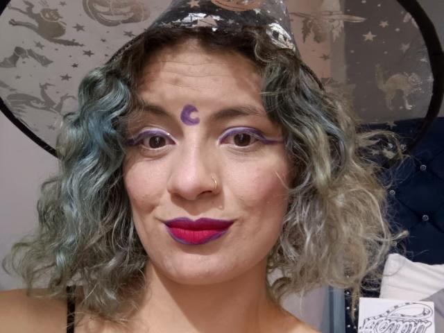 IndiGoth has curly blue-gray hair, purple eyeliner, a crescent moon tattoo on her forehead, purple lipstick, and a silver nose ring, wearing a star-patterned hat.