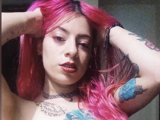 ErikaValeska with pink hair, nose ring, tattoos, choker, and bracelet, arms raised, showing tattoos on chest and arm, and blue floral tattoo on right arm.