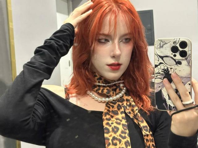 MissVelora with red hair, black long-sleeve shirt, leopard scarf, pearl necklace, red lipstick, holding phone in mirror, hand in hair, wearing ring, smartphone case with anime art.