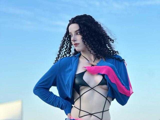 MissVelora with curly black hair, blue jacket, black strappy bikini, pink gloves, fair skin, looking to the side, hand on chest, showing small tits, against a blue sky.