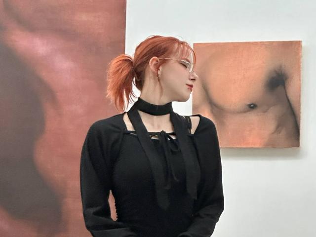 MissVelora has red hair in a ponytail, wears glasses, a black choker, and a black dress with cutouts. She stands in front of a nude torso painting.