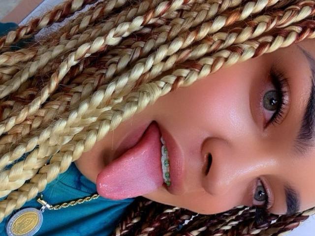 KarinaWine with blonde and brown braided hair, blue eyes, and her tongue out, showing her green braces. Her pink tongue is slightly wet and her face is close.