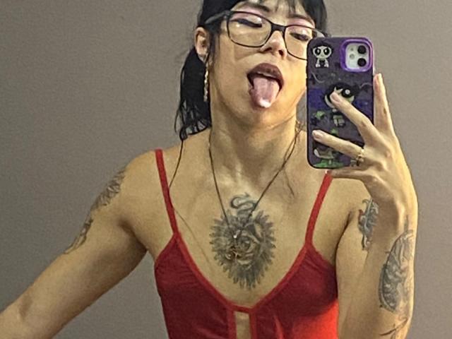 MiaDeLune, avec ses cheveux noirs, ses lunettes et ses tatouages, la langue tirée, portant une robe rouge en spaghetti, tenant un téléphone avec une coque à motif de crâne, seins visibles, tatouage sur la poitrine et piercings aux mamelons..