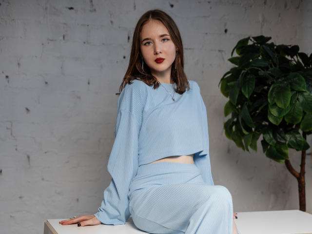 MarieLoving with pale skin and brown hair, wearing light blue crop top and matching skirt, sitting on white table, small tits, red lipstick, green plant to the right.