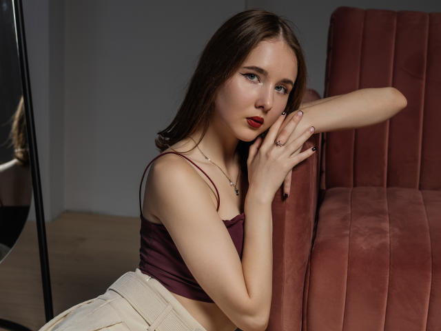 MarieLoving has pale skin, brown hair, red lipstick, and is wearing a purple crop top and beige skirt, leaning on a pink velvet chair with one arm.