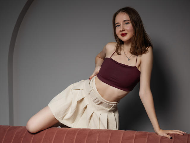 MarieLoving leans on a pink velvet couch. She has fair skin, brown hair, and wears a maroon strapless crop top and a beige pleated skirt. Her right hand rests on the couch while her left is on her hip. She has red lipstick and a small necklace with a cross. The image is sharp with a neutral gray background. Her body is angled to the right, and she looks at the camera with a slight smile.