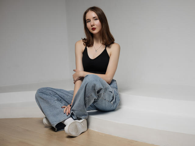 MarieLoving, with small tits, sitting cross-legged, wearing a black tank top, blue jeans, and white sneakers, has brown hair, red lipstick, and a necklace, in a minimalistic room.